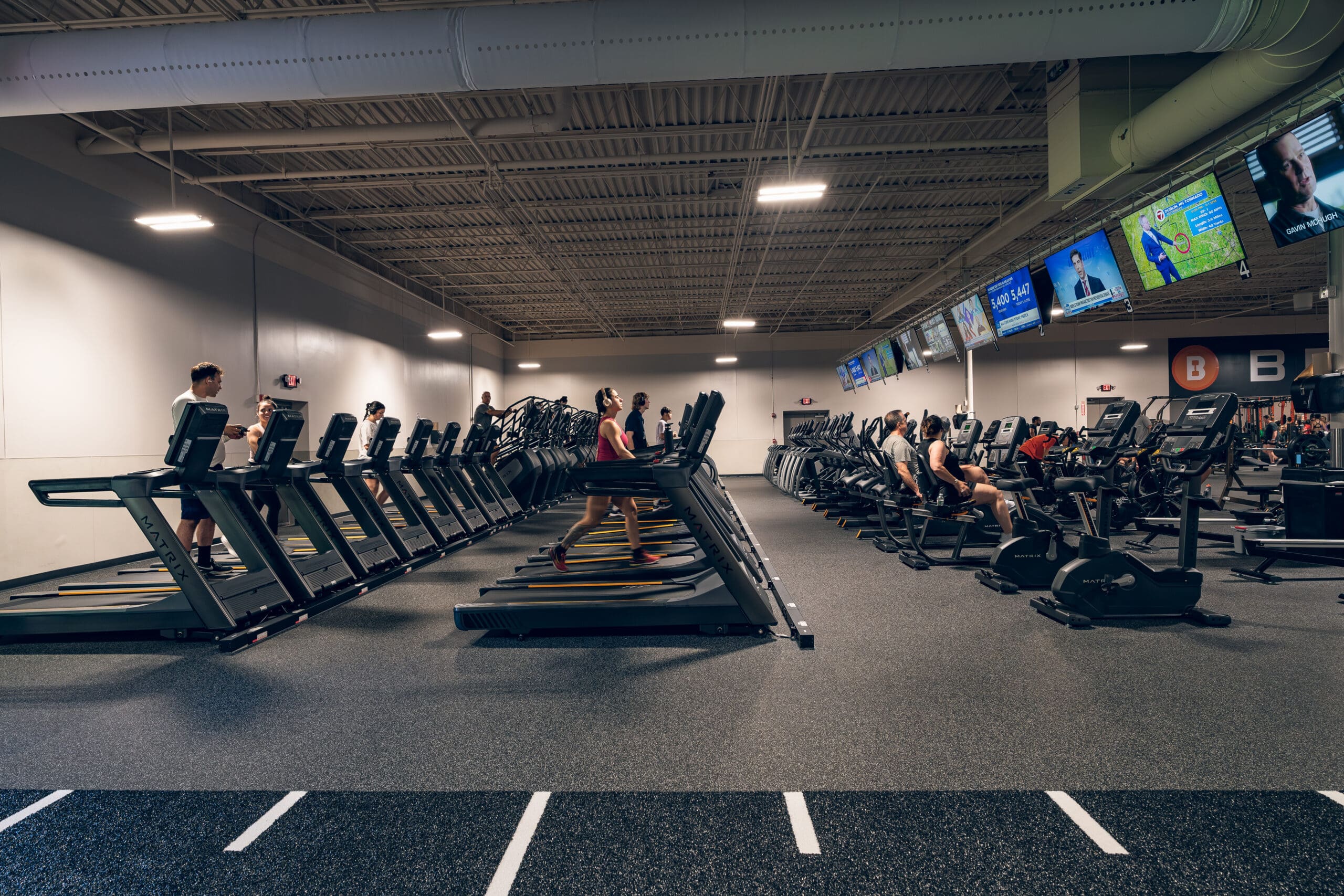 Rows of treadmills and cardio machines at Best Fitness Woburn with members exercising and TVs overhead.
