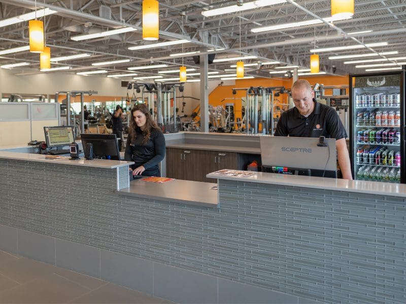 2 gym team members sit at the front desk in the lobby of a modern best fitness gym ready to greet guests as they come in